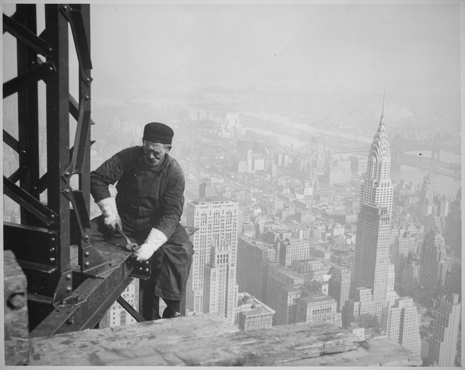 Operário de boné sentado numa viga, apertando uma peça, com Manhattan e o Chrysler Building lá embaixo