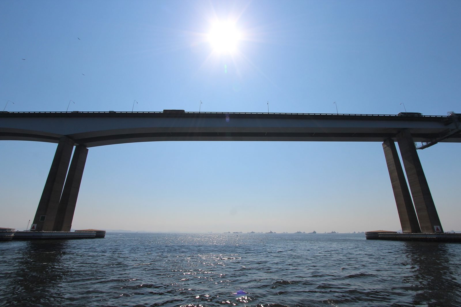 O vão central de aço da Ponte Rio–Niterói visto de baixo, a partir da água, com o sol ao fundo