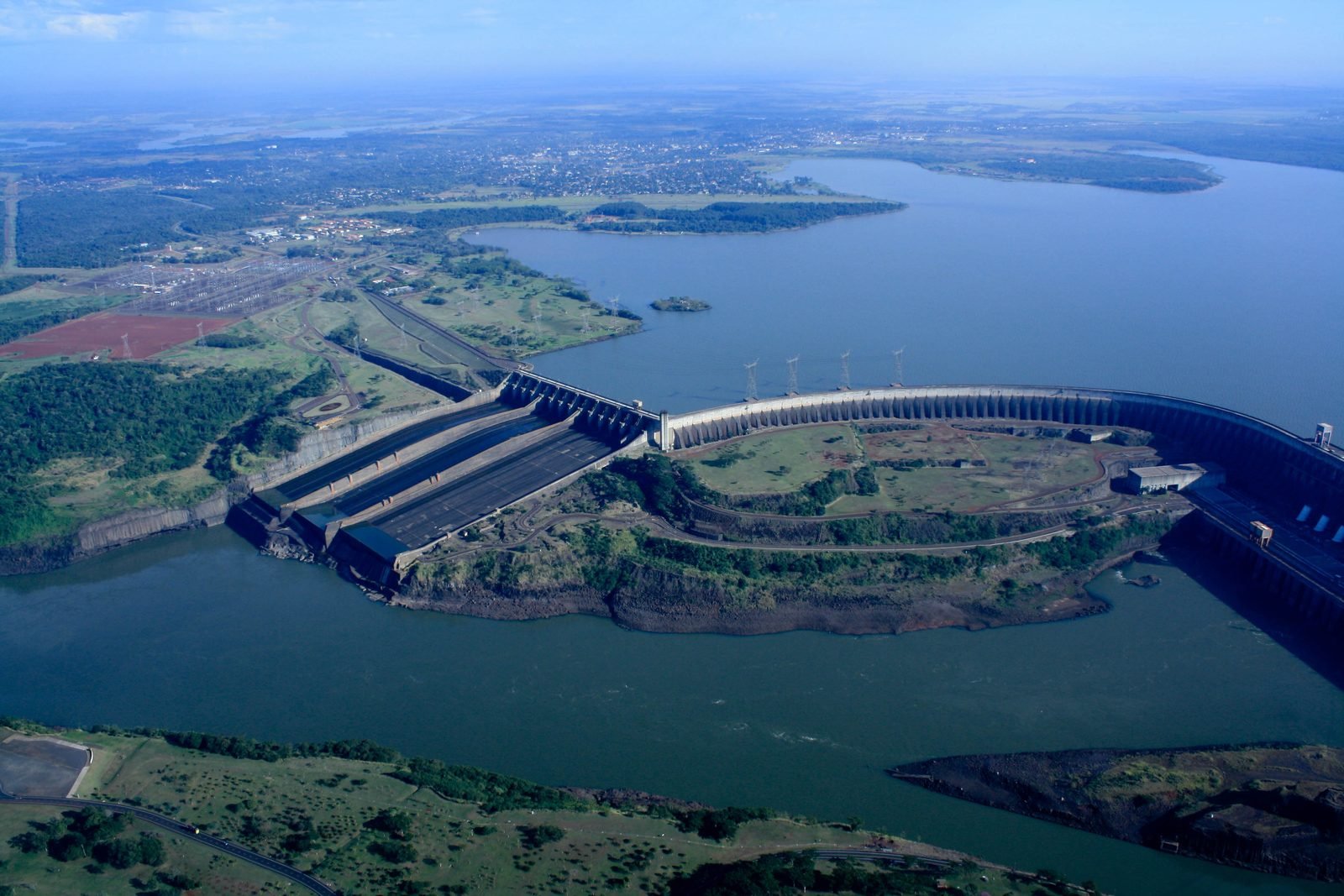 Aerial view of the Itaipu Dam: the spillway on the left, the curving dam and the reservoir behind, with the Paraná River in the foreground