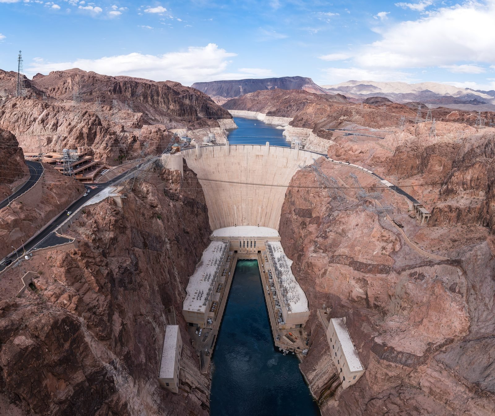 Hoover Dam seen from above, with the curved concrete wall between the rocks of Black Canyon, the power plant at its foot and Lake Mead in the background