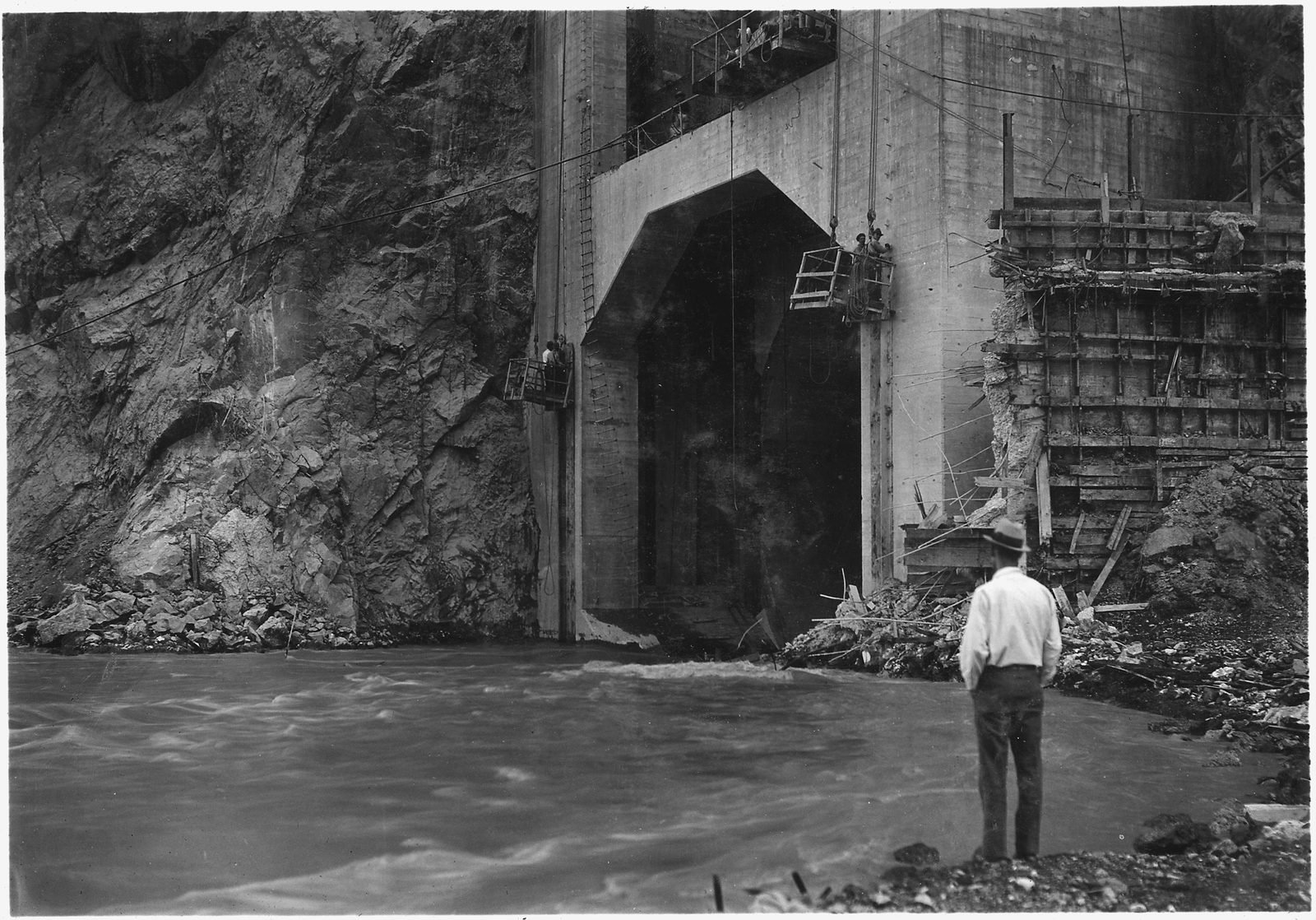Agua del río saliendo por un portal de túnel de concreto en la pared del cañón, con un hombre de camisa blanca mirando de cerca