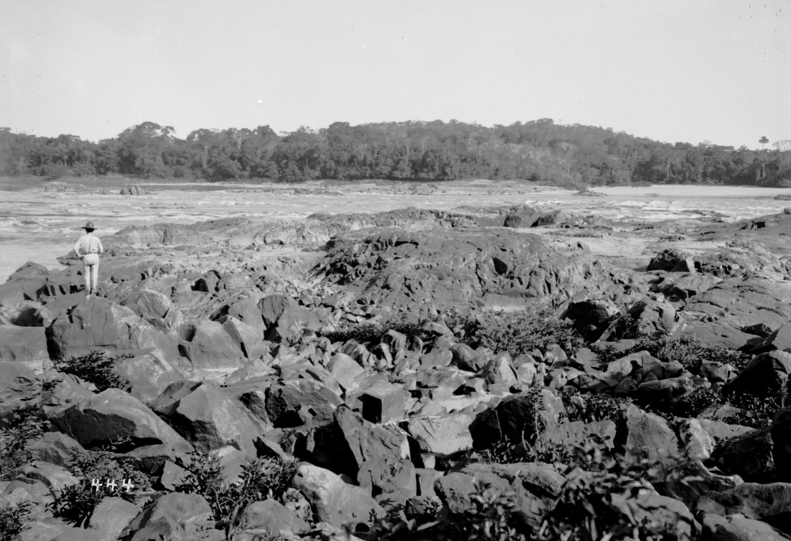 Riverbed covered with large blocks of dark rock, with a man standing among the stones and forest on the far bank