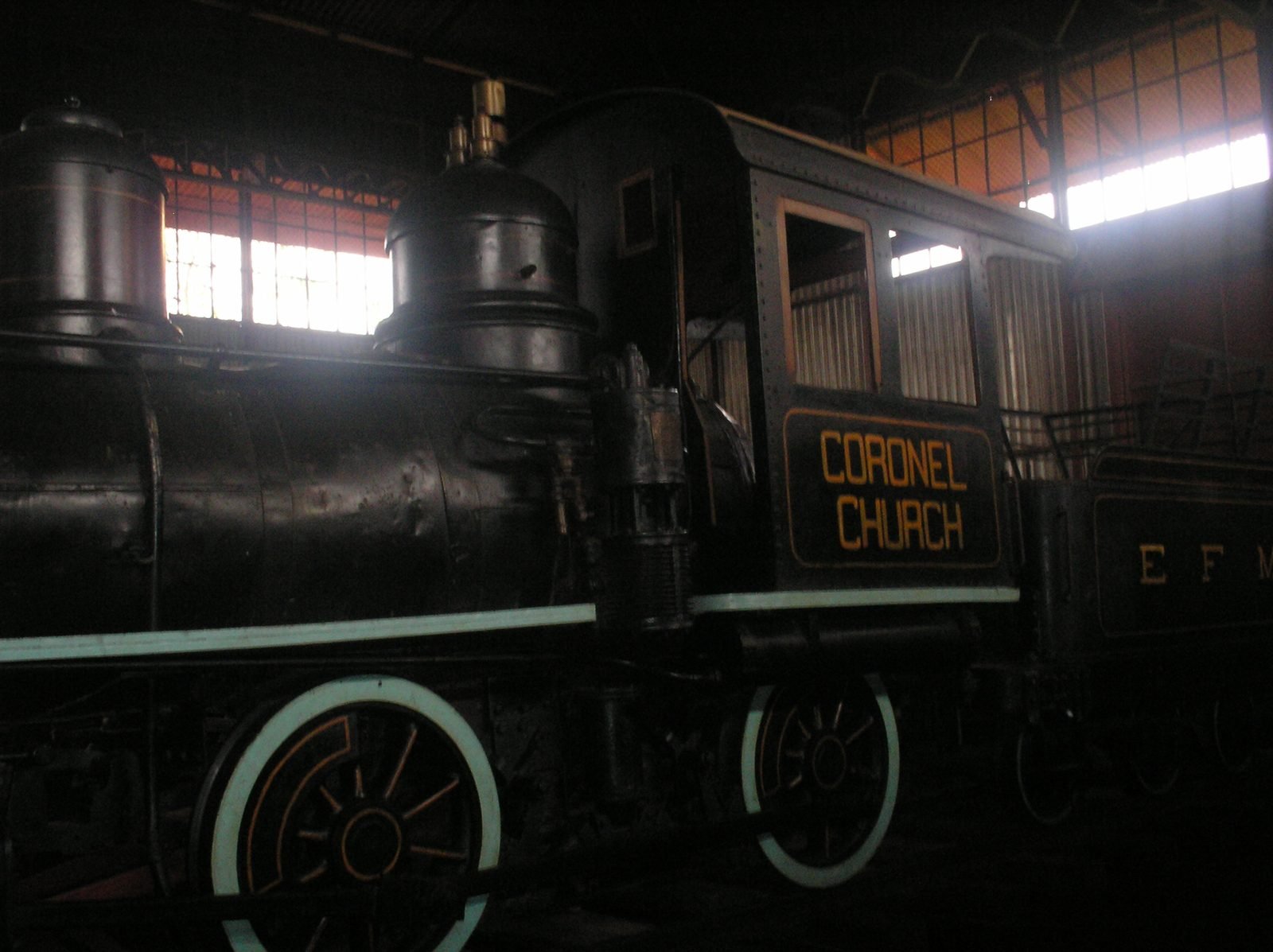 Black steam locomotive with pale-rimmed wheels and the name Coronel Church on the cab, inside a shed