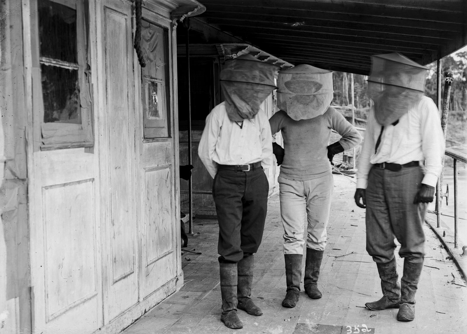 Three men in boots on a covered wooden porch, their heads wrapped in mesh hoods