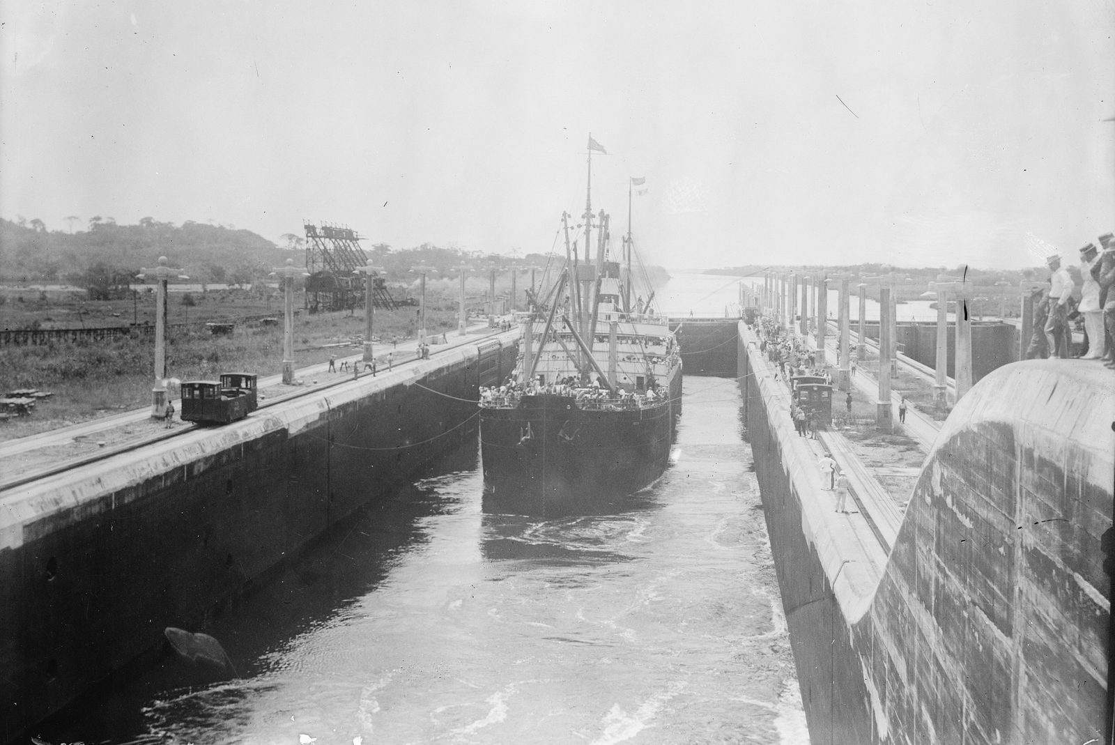 Navio a vapor com passageiros no convés dentro de uma câmara de eclusa estreita, com pequenas locomotivas nos muros laterais e pessoas observando