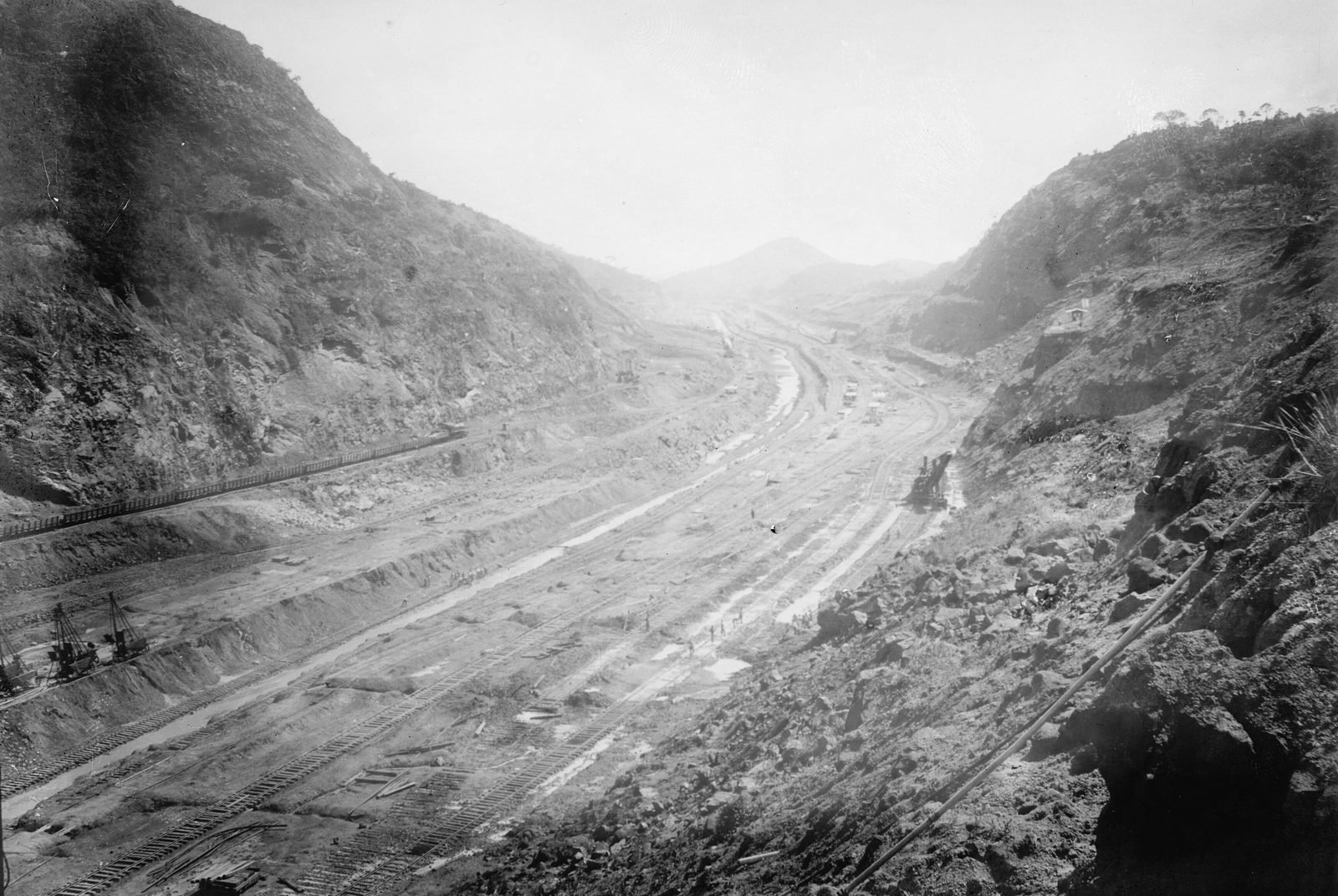Deep, dry valley cut between two rock slopes, with several railroad tracks along the bottom, a steam shovel on the right and cranes on the left