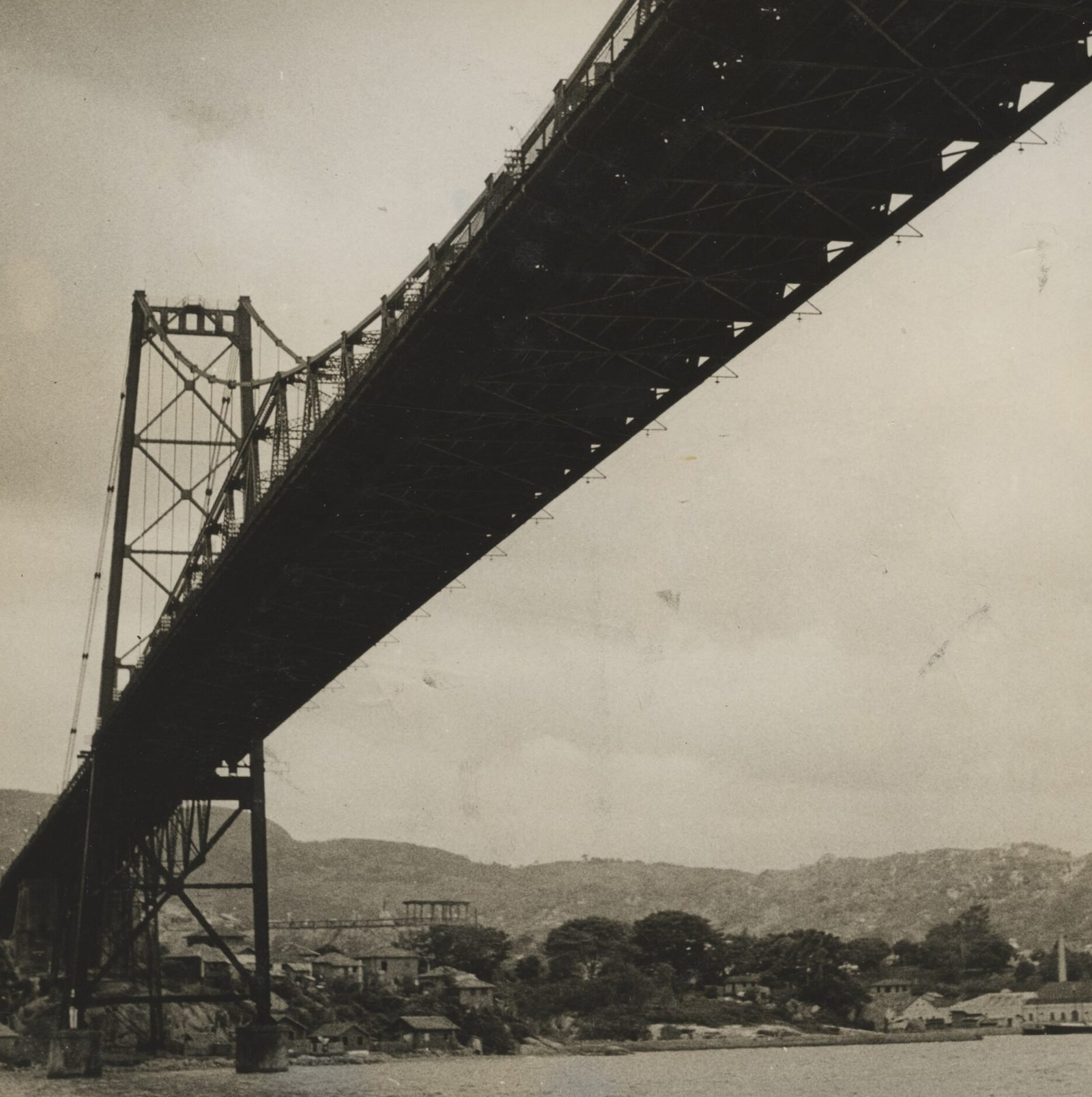 Fotografia antiga em preto e branco: a ponte vista de baixo, do mar, com o tabuleiro treliçado escuro cruzando o céu na diagonal e uma torre à esquerda; casas e morros ao fundo