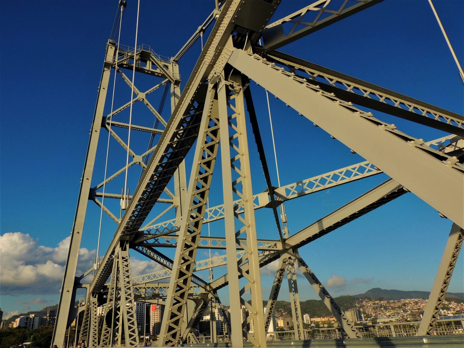 Vista de baixo para cima de barras de aço cinza cruzadas, cheias de rebites, com uma torre treliçada e pendurais contra o céu azul