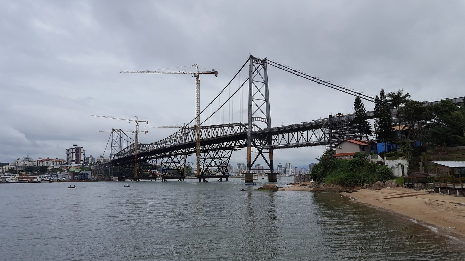A ponte sob céu nublado com guindastes de torre amarelos ao lado do vão central e estruturas de apoio provisórias sob o tabuleiro