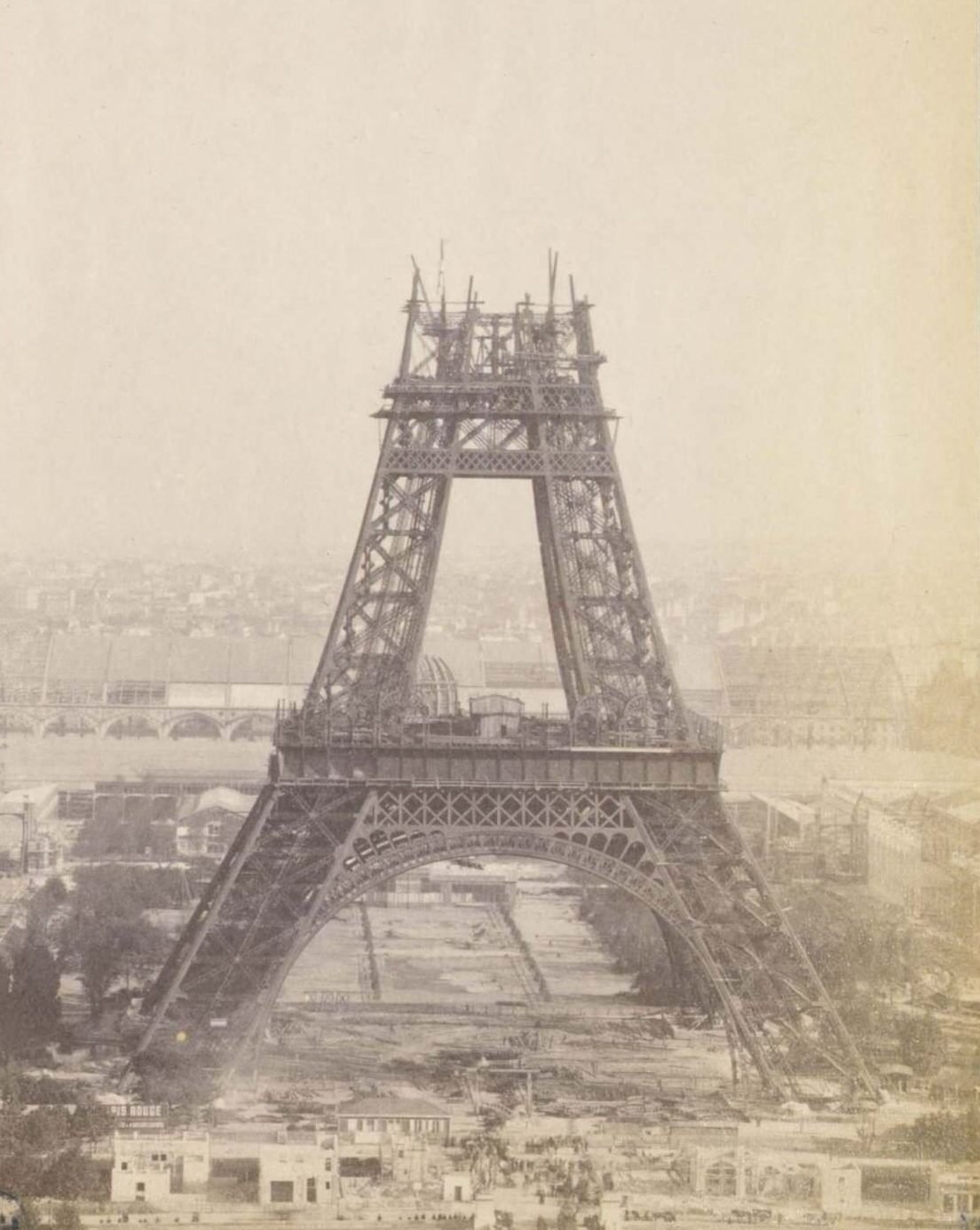 Fotografia antiga em sépia: a torre até um pouco acima do segundo andar, com o arco da base e o primeiro andar completos e gruas no topo inacabado, sobre o Champ-de-Mars em obras e a cidade ao fundo na névoa