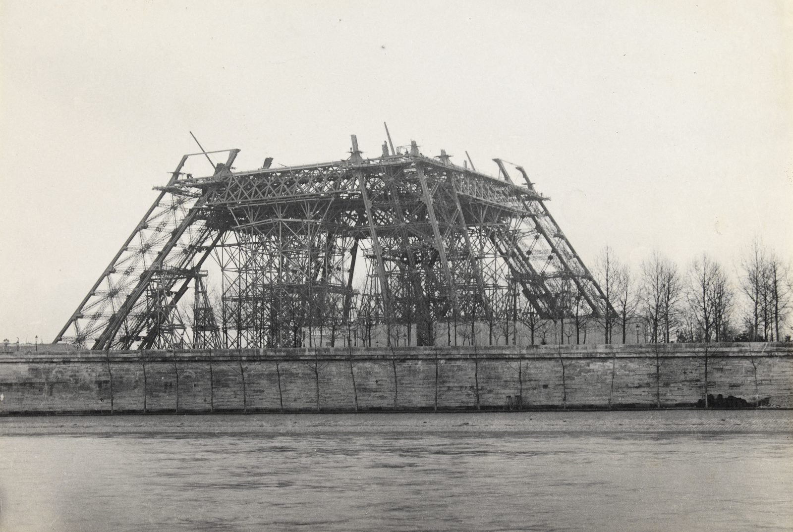 Old black-and-white photograph: the four sloping legs of the tower, already joined by the first-floor structure, resting on a forest of wooden scaffolding, behind the Seine quay wall and leafless trees