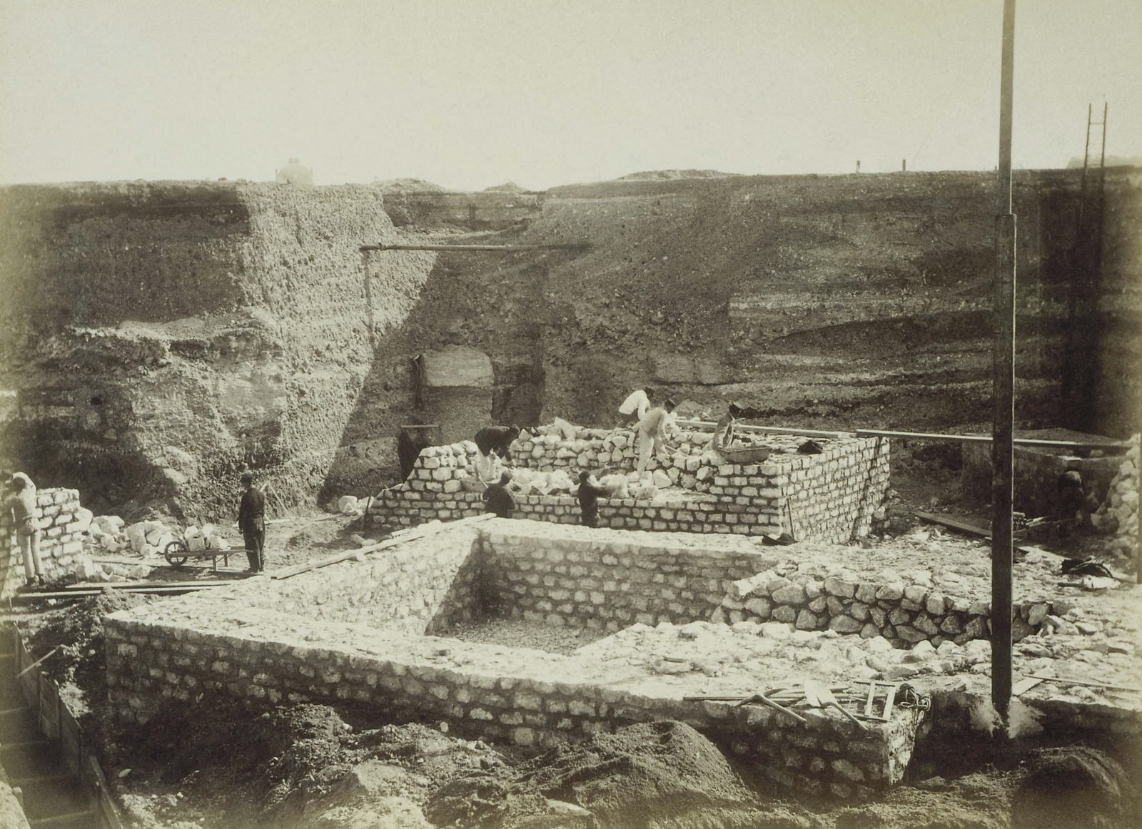Fotografia antiga em sépia de uma escavação funda com paredes de terra, onde operários de chapéu erguem blocos retangulares de alvenaria de pedra; um carrinho de mão e um poste de madeira em primeiro plano