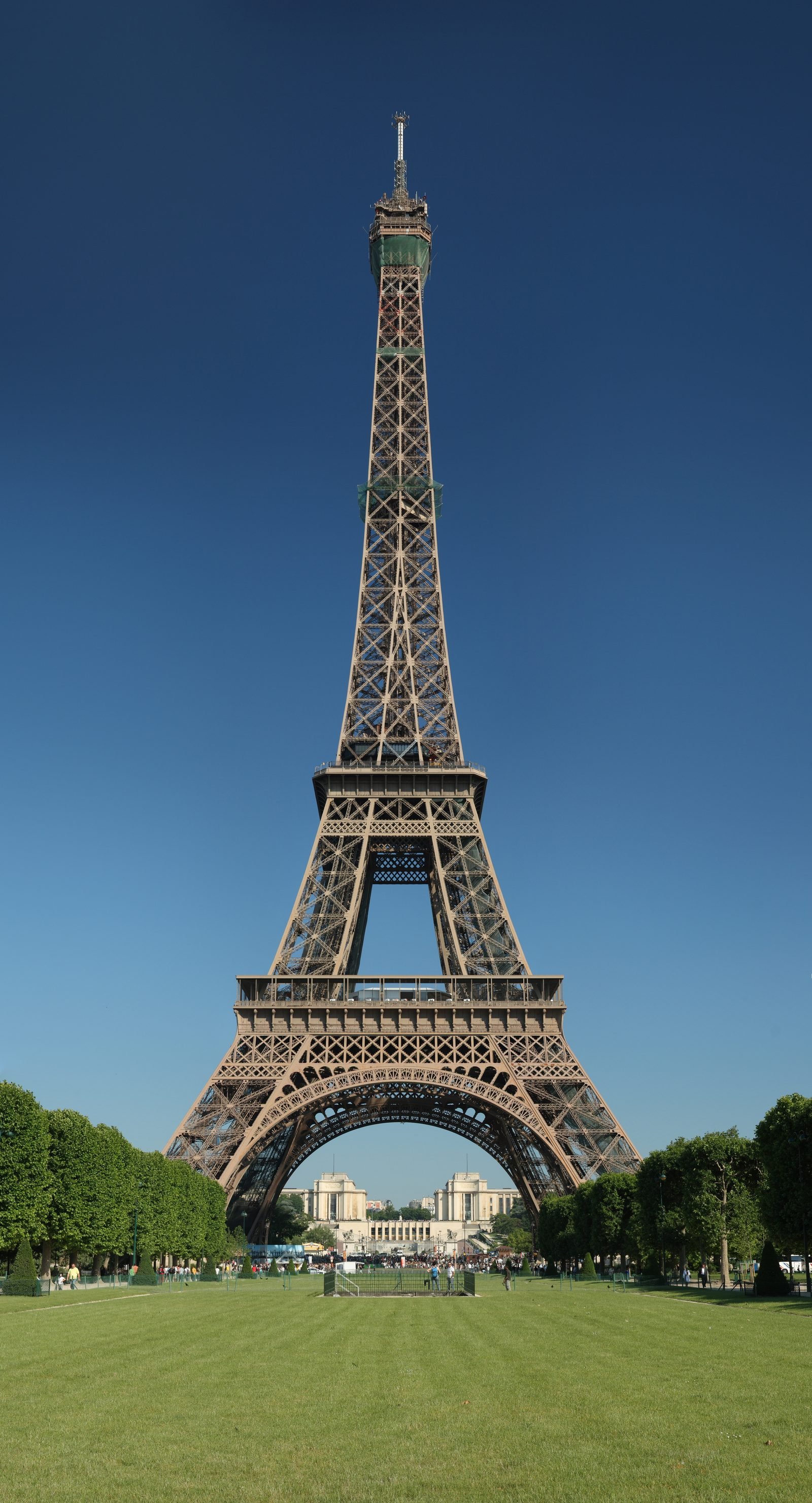 A torre vista de frente, de dia, contra o céu azul, com o gramado do Champ-de-Mars em primeiro plano e árvores dos dois lados