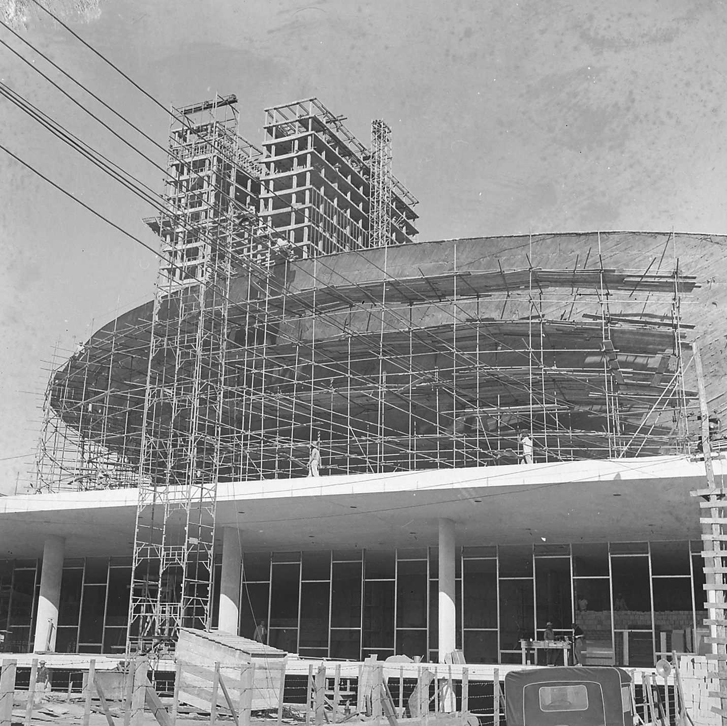 Old black-and-white photograph: the concave dome of the Chamber covered in tubular scaffolding, on the platform slab supported by pilotis with a glass facade; behind it, the skeleton of one of the towers under construction