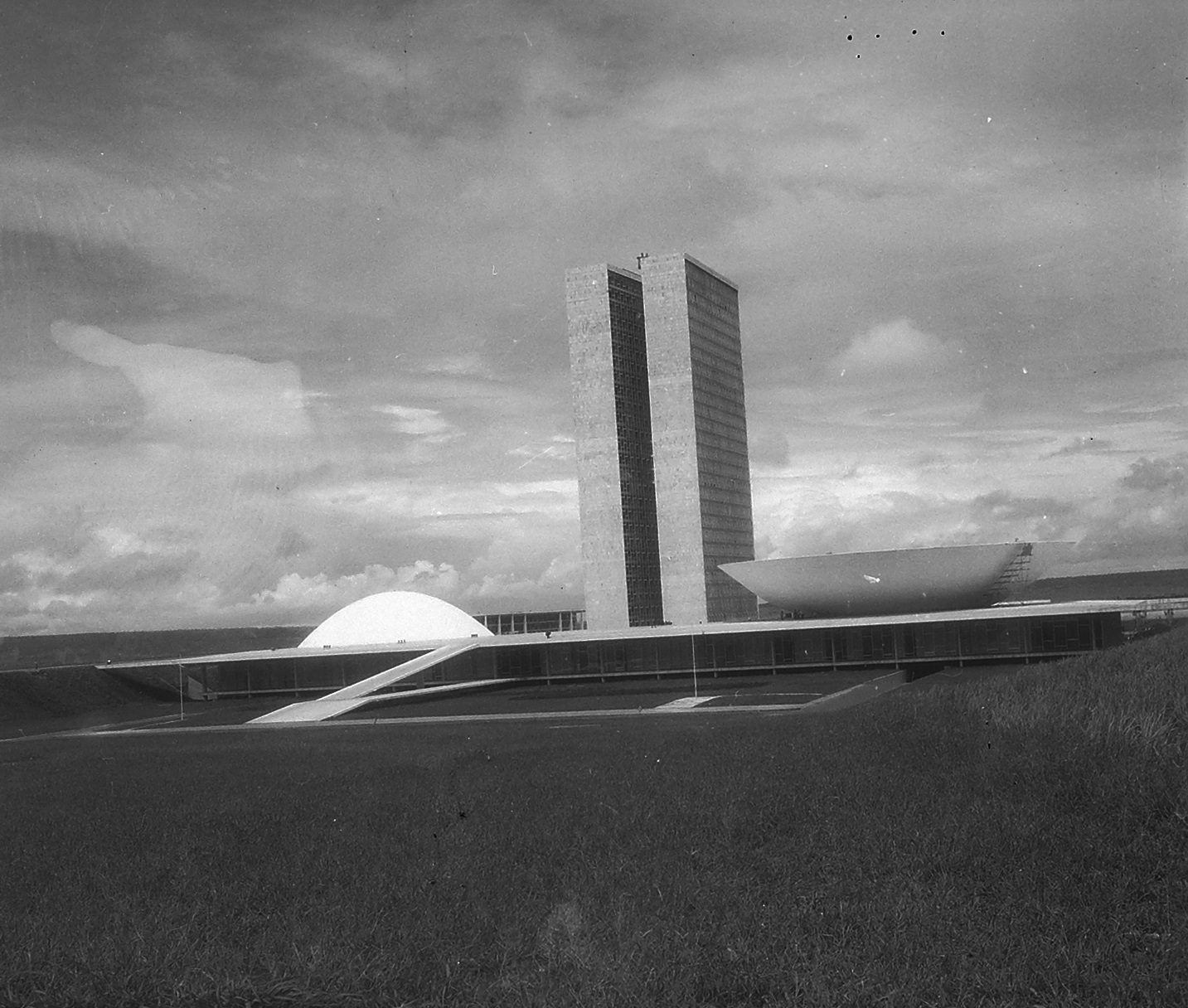 Old black-and-white photograph of the complex seen from a distance, across a lawn: the convex dome on the left, the two towers in the center, the concave dome on the right and the ramp rising to the platform
