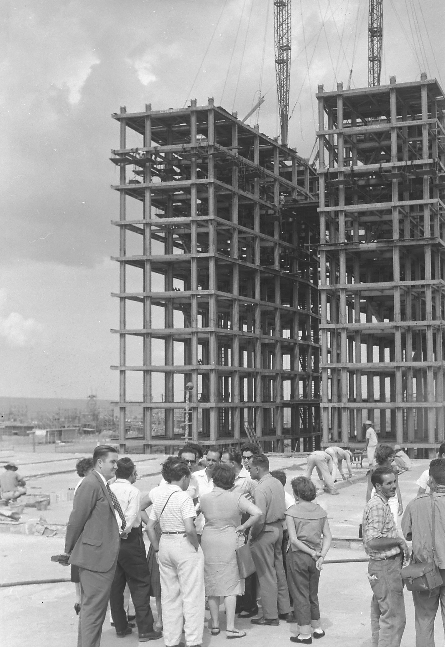 Old black-and-white photograph: the skeleton of columns and beams of the two Congress towers, still without walls, with cranes at the top, and a group of visitors in suits and dresses on the site in the foreground