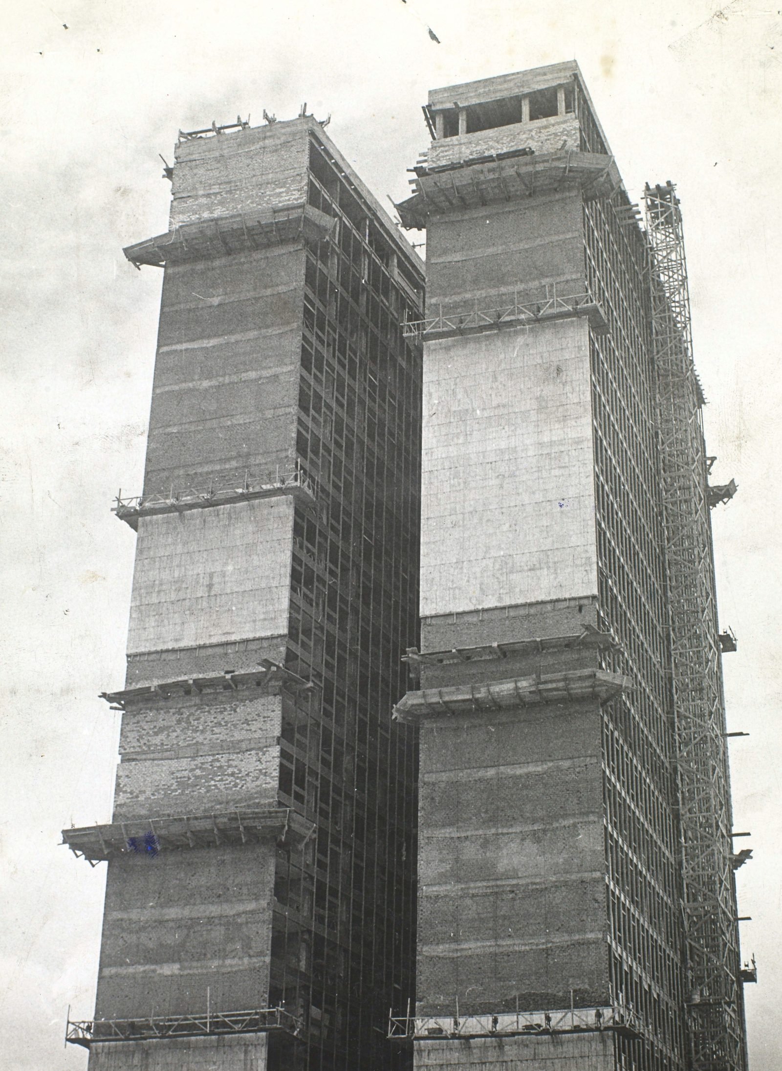 Old black-and-white photograph: the two towers seen from below, already clad at the ends, with scaffolding platforms hanging on several floors, windows without glass and a crane tower alongside on the right