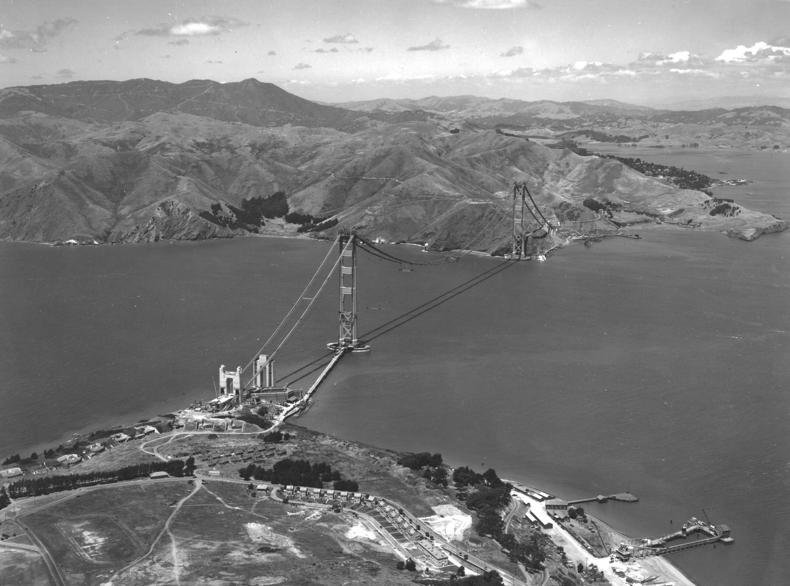 Fotografia aérea em preto e branco: duas torres de aço sobre o estreito ligadas por dois cabos em curva, sem tabuleiro entre elas, com morros ao fundo e casas em primeiro plano