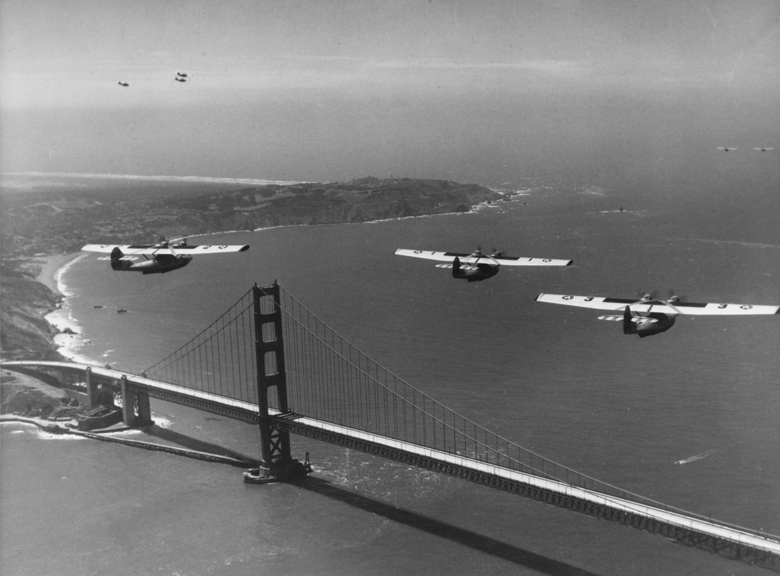 Fotografia em preto e branco: três hidroaviões em formação sobre uma ponte pênsil pronta, com uma torre, o tabuleiro inteiro e a costa ao fundo