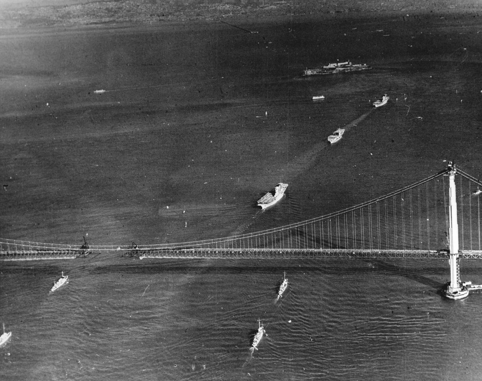 Fotografia aérea em preto e branco: navios de guerra em fila passando sob uma ponte pênsil cujo tabuleiro ainda tem uma abertura no meio; uma torre à direita