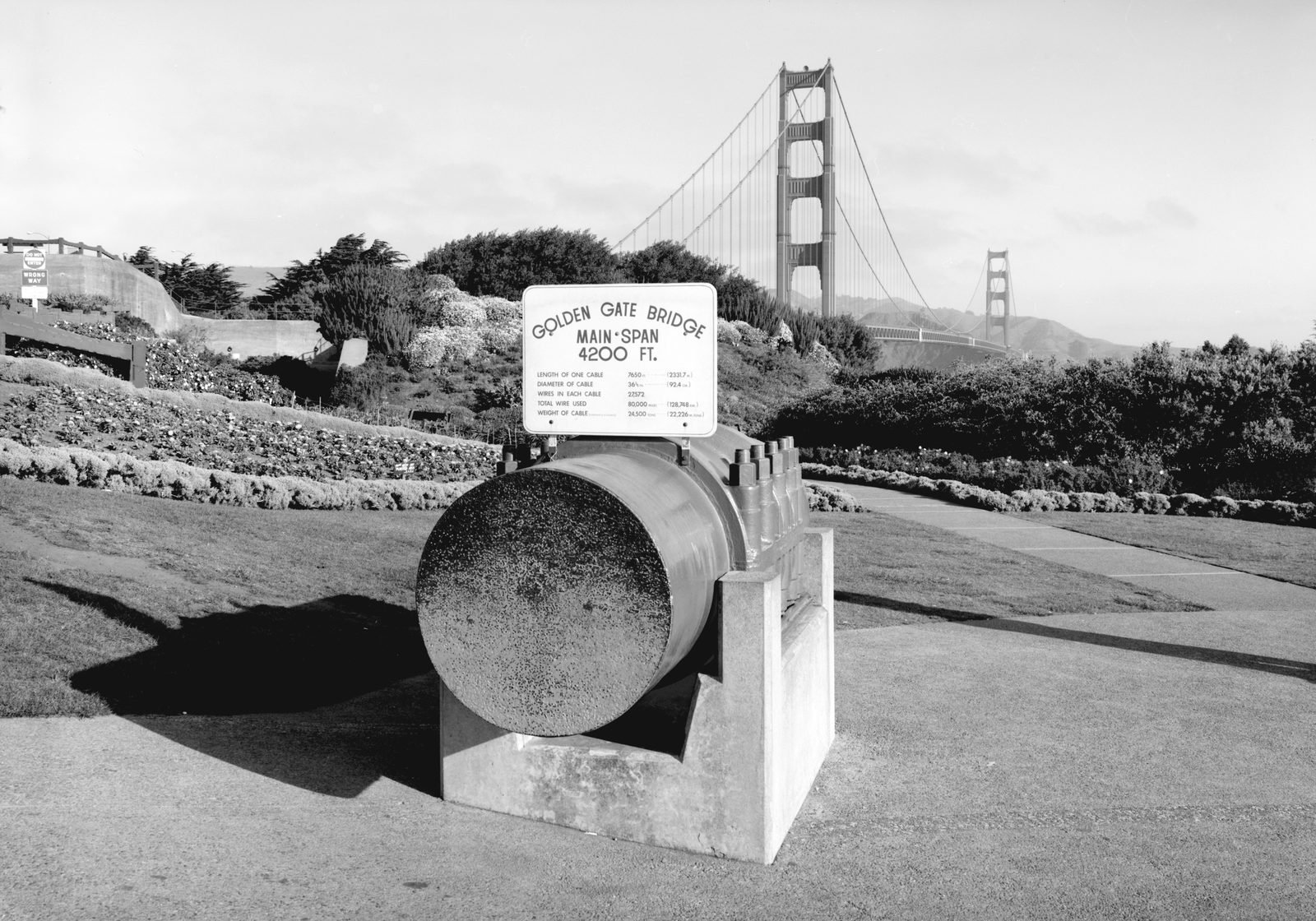 Fotografia em preto e branco de um grande cilindro metálico exposto num jardim, com uma placa sobre ele com os dados do cabo; a ponte e suas duas torres ao fundo