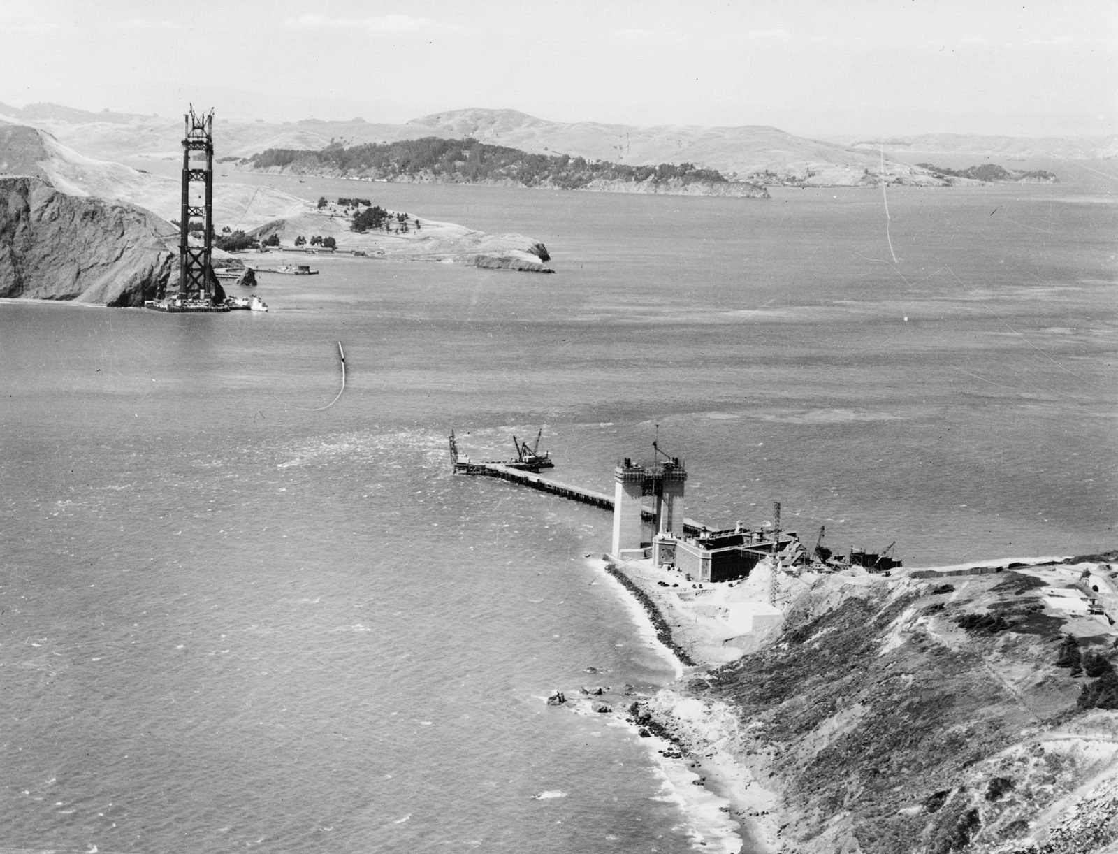 Fotografia aérea antiga em preto e branco: ao fundo, uma torre de aço solitária junto a um penhasco do outro lado do estreito; em primeiro plano, uma passarela estreita avança da praia para o mar até guindastes e blocos de concreto