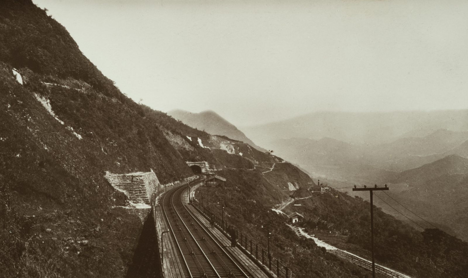 Old sepia photograph: a double-track railway curves along a steep slope, with a stone wall on the left, a tunnel in the background, telegraph poles and the mist-covered valley on the right