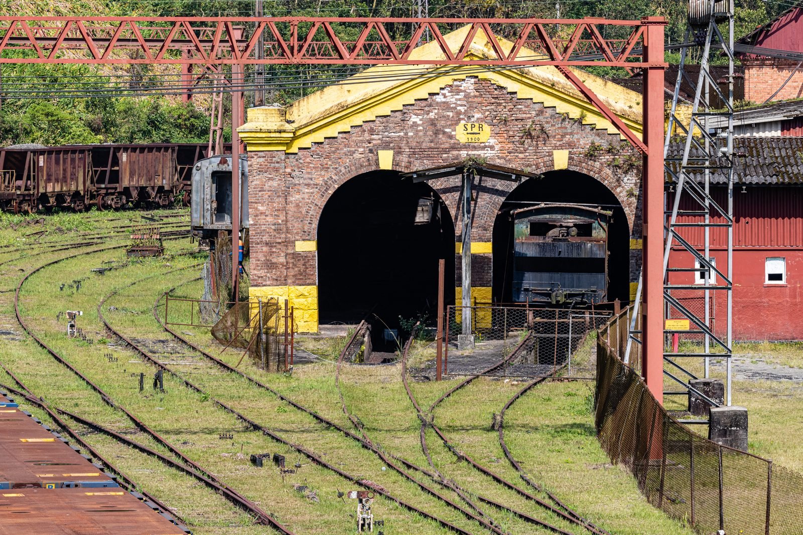 Galpão de tijolos com dois arcos e frontão amarelo com a inscrição SPR 1900, um vagão antigo dentro de um dos arcos, trilhos cobertos de grama na frente e pórticos metálicos vermelhos