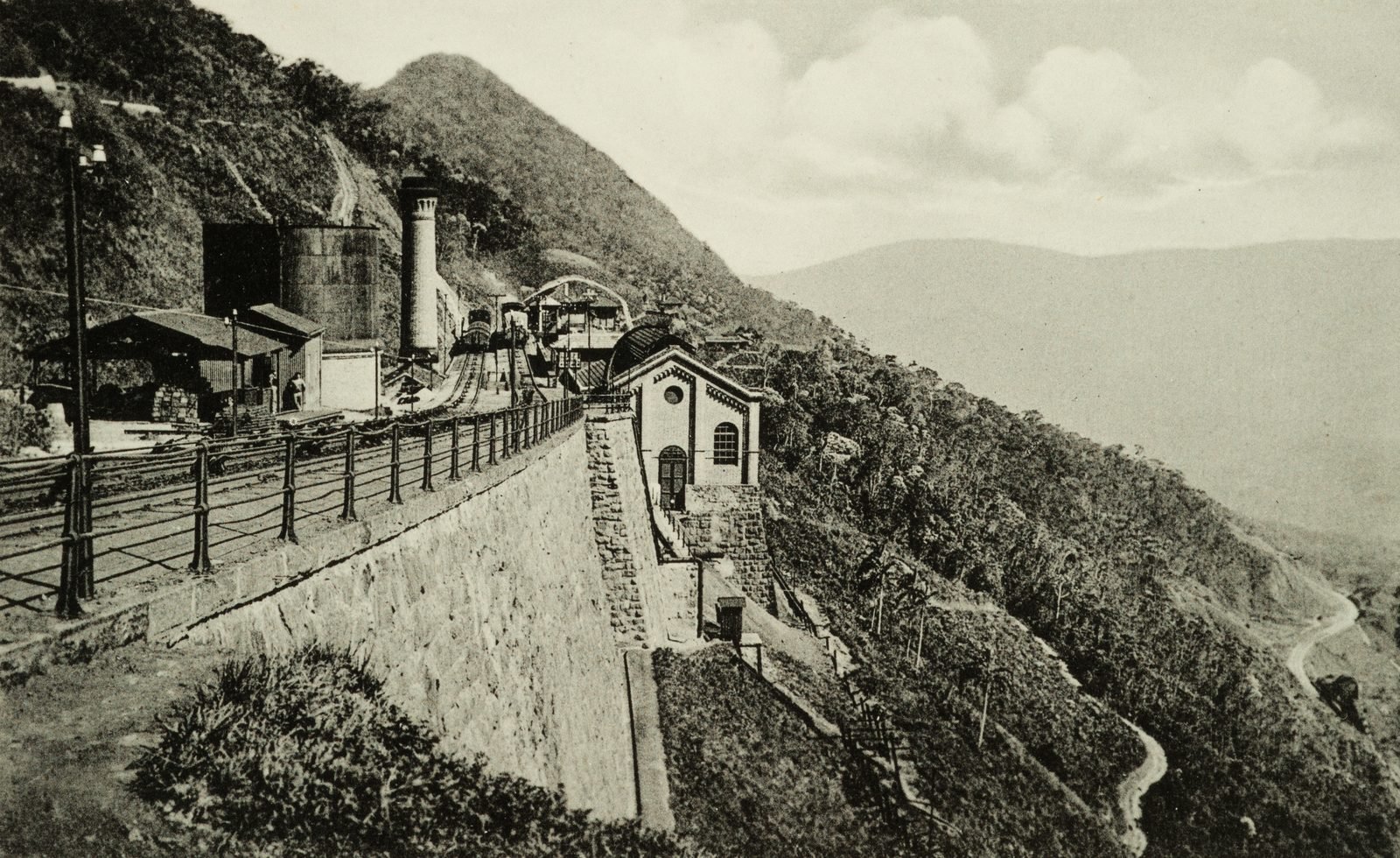 Cartão-postal antigo em preto e branco: no alto de uma encosta, trilhos com grade de ferro sobre um muro alto de pedra, uma chaminé, tanques e uma pequena casa de alvenaria, com a serra e o vale ao fundo