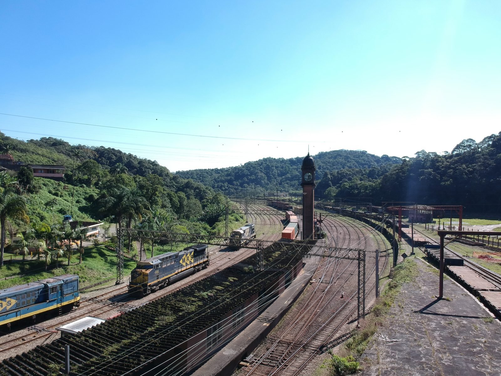 Vista aérea de um pátio ferroviário entre morros de mata, com muitas linhas, locomotivas azuis e amarelas, uma torre de relógio de tijolo no centro e plataformas vazias à direita