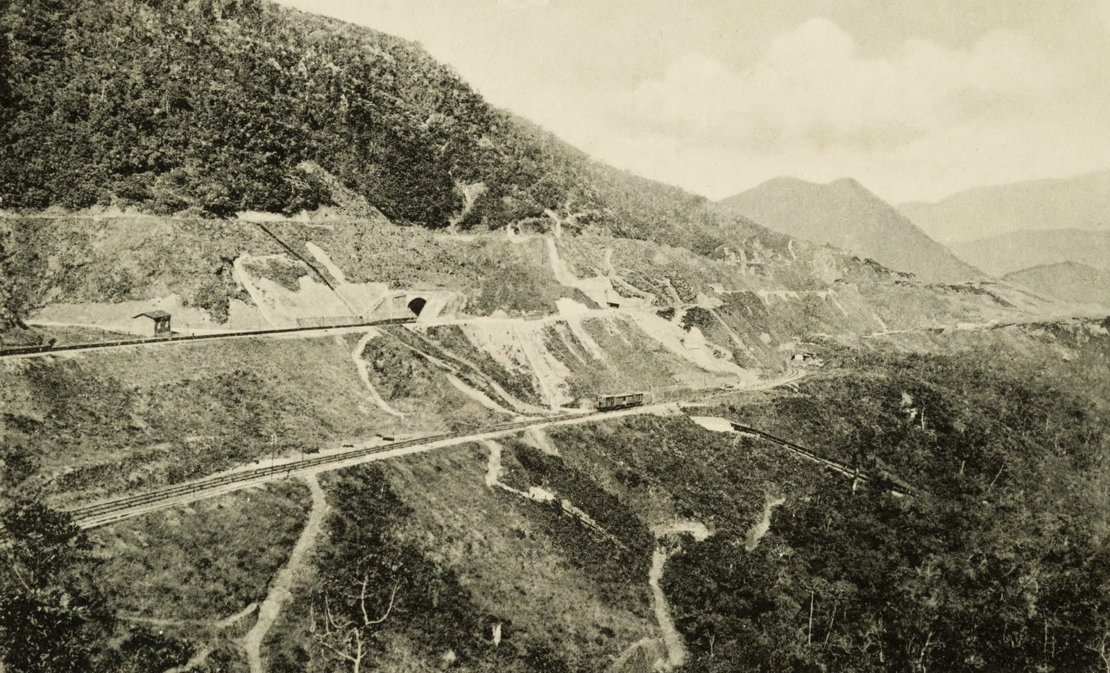 Cartão-postal antigo em preto e branco: vista do alto de uma encosta desmatada, com duas linhas férreas cortadas em níveis diferentes, a boca de um túnel e um vagão pequeno no meio da rampa