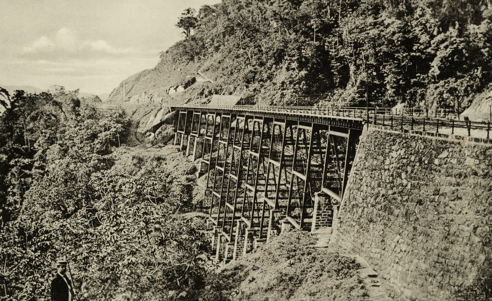 Cartão-postal antigo em preto e branco: um viaduto de ferro sobre pilares treliçados atravessa um vale de mata, apoiado à direita num grande muro de pedra; um homem de quepe aparece no canto inferior esquerdo