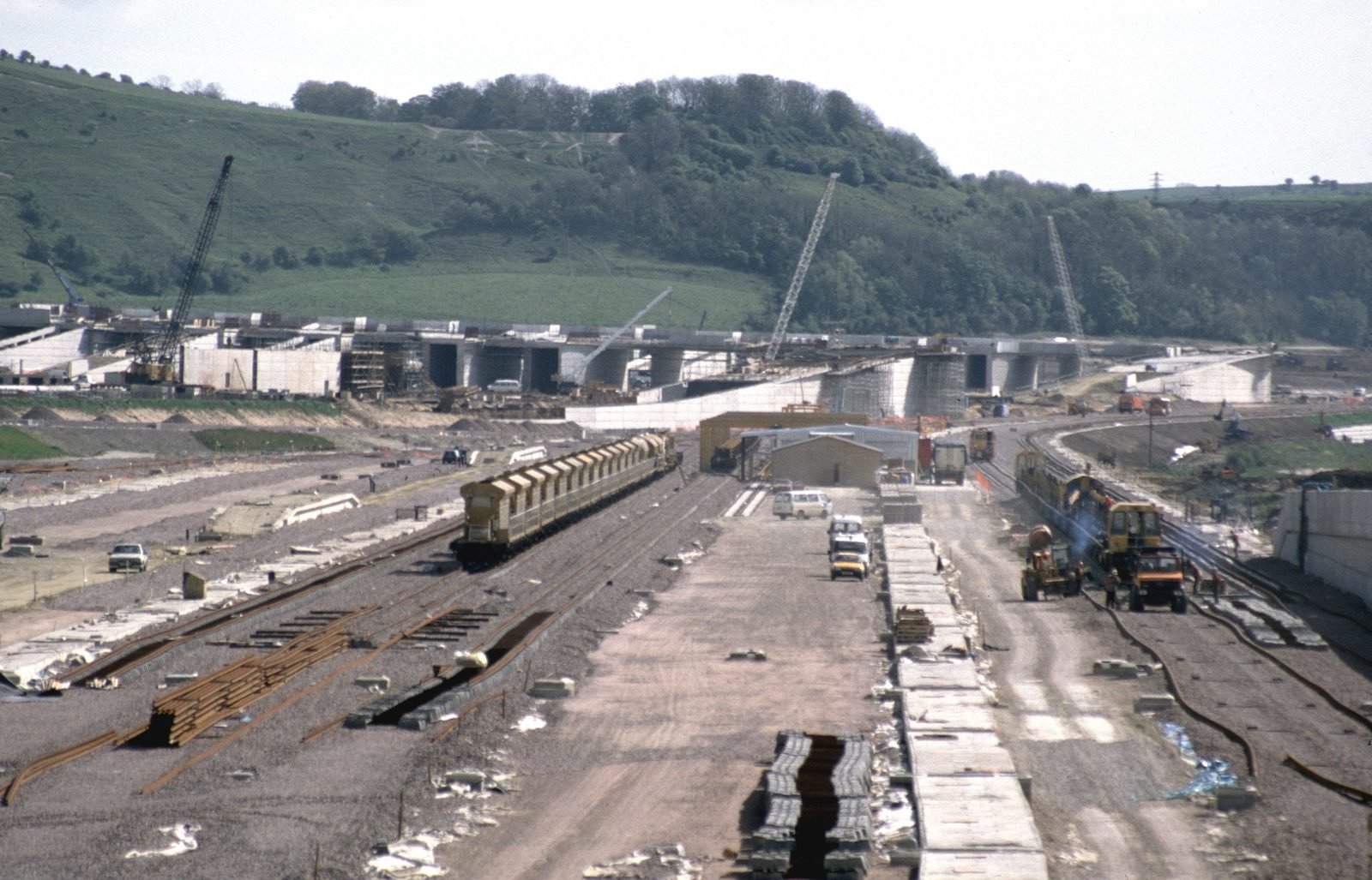Wide construction site with tracks, stacks of sleepers, wagons and work locomotives, concrete viaducts under construction and cranes in front of a green hill