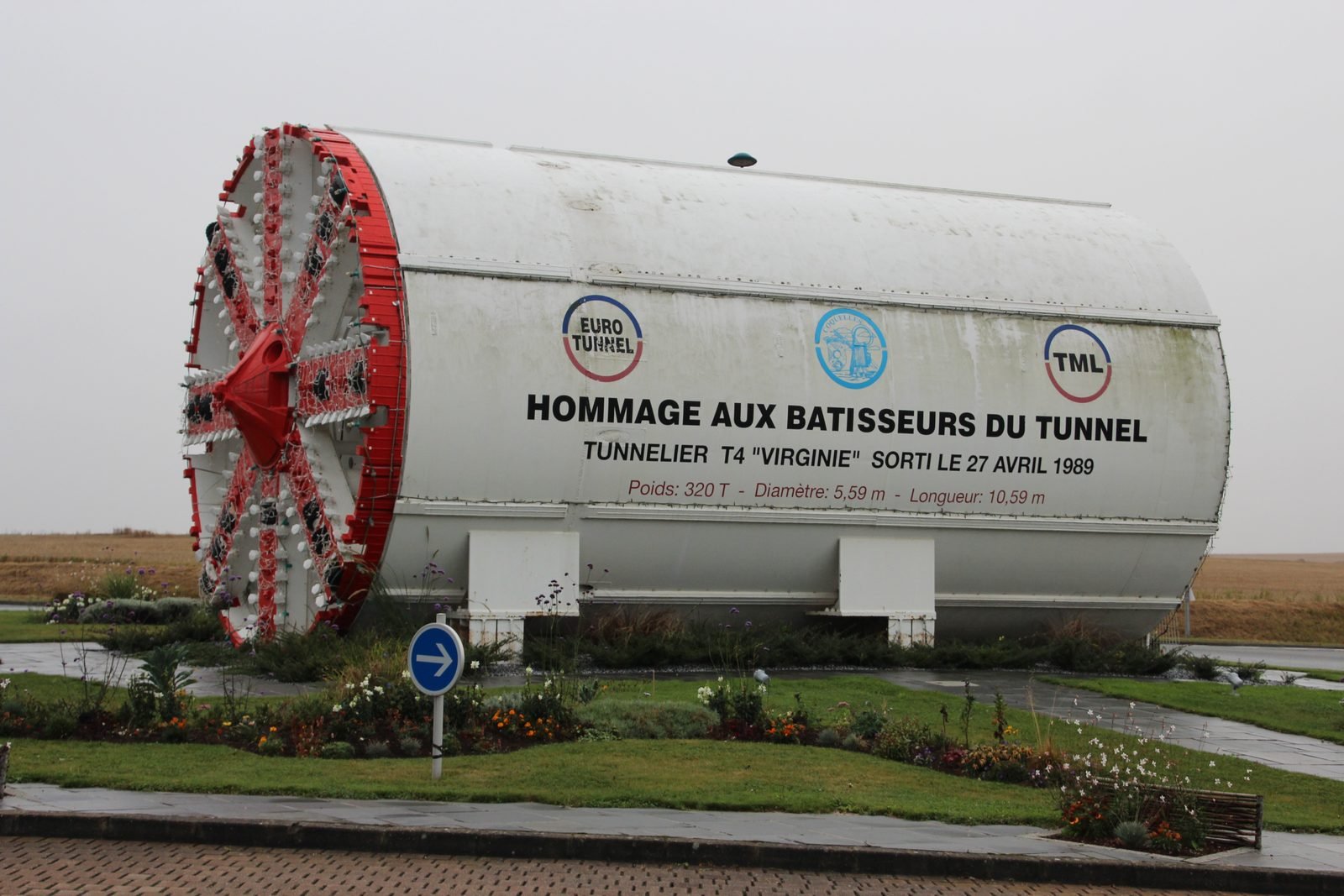 White cylindrical tunnel boring machine displayed in a roundabout garden, with the red wheel-shaped cutting head on the left and the French inscription Hommage aux bâtisseurs du tunnel on its body