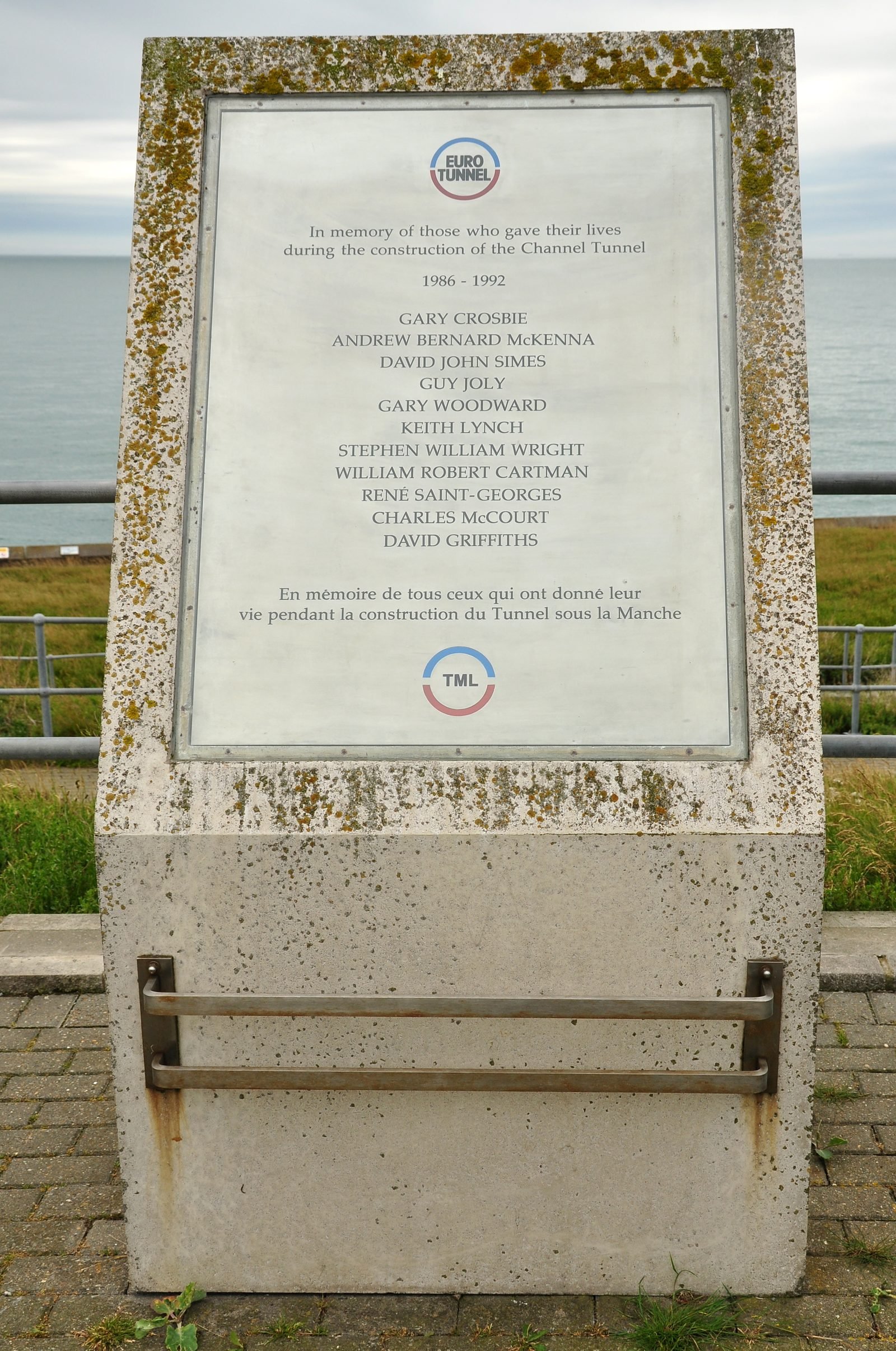 Metal plaque on a concrete block by the sea, with text in English and French in memory of those who died building the tunnel and a list of eleven names