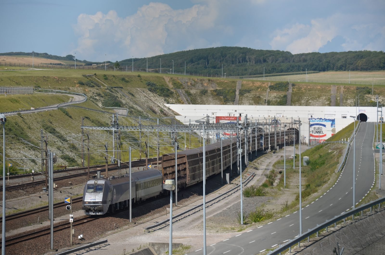 Silver train hauling truck-carrying wagons emerges from a white concrete portal cut into a hillside, under catenary wires, with a curving road on the right