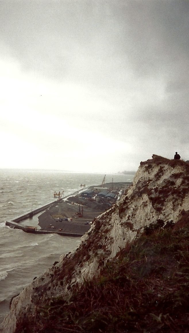 View from the top of a white cliff over the gray sea: below, a platform of earth enclosed by a dam juts out over the water, with blue sheds and cranes