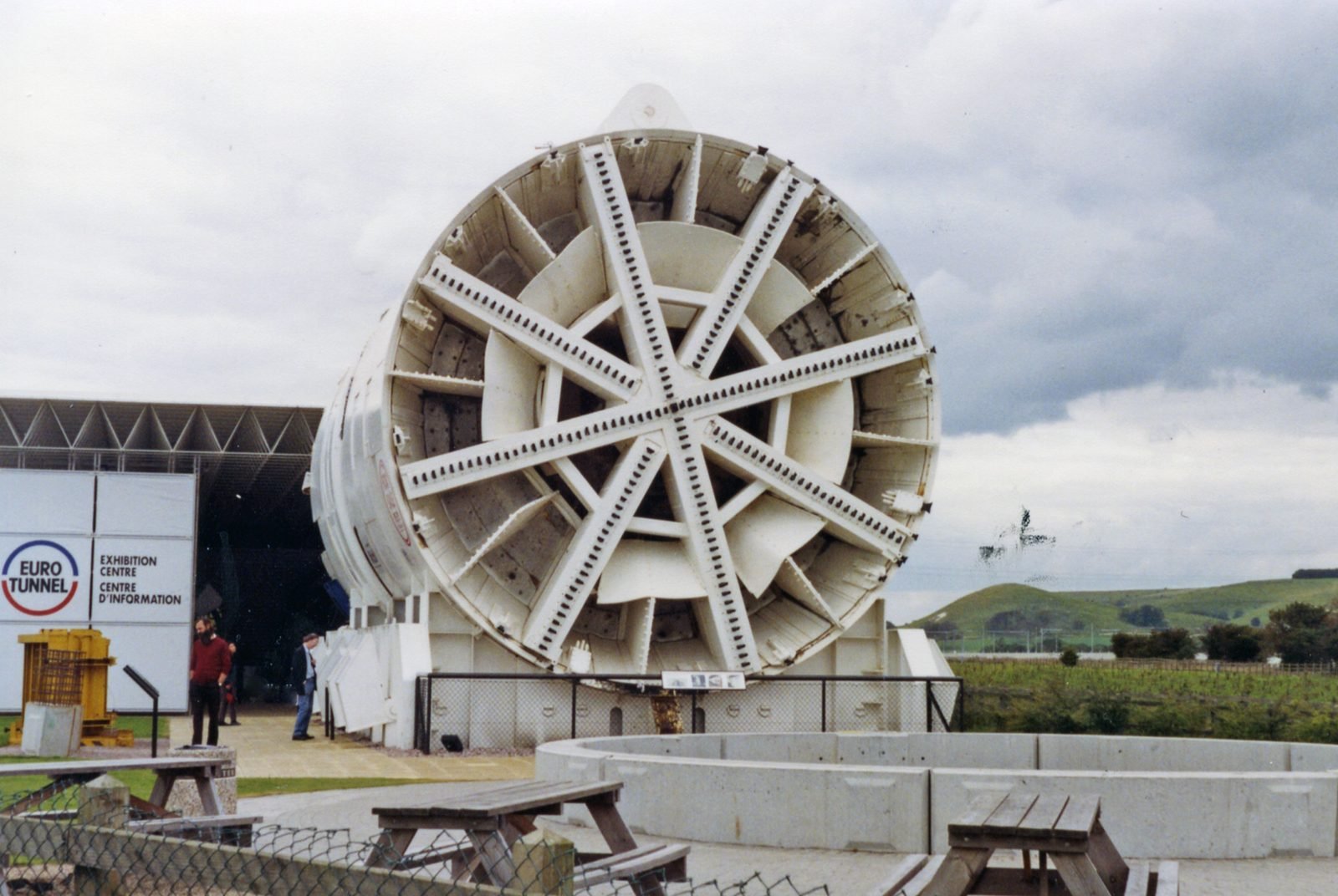 Circular cutting head of a tunnel boring machine, with radial arms full of teeth, displayed outdoors next to a pavilion bearing the Eurotunnel logo