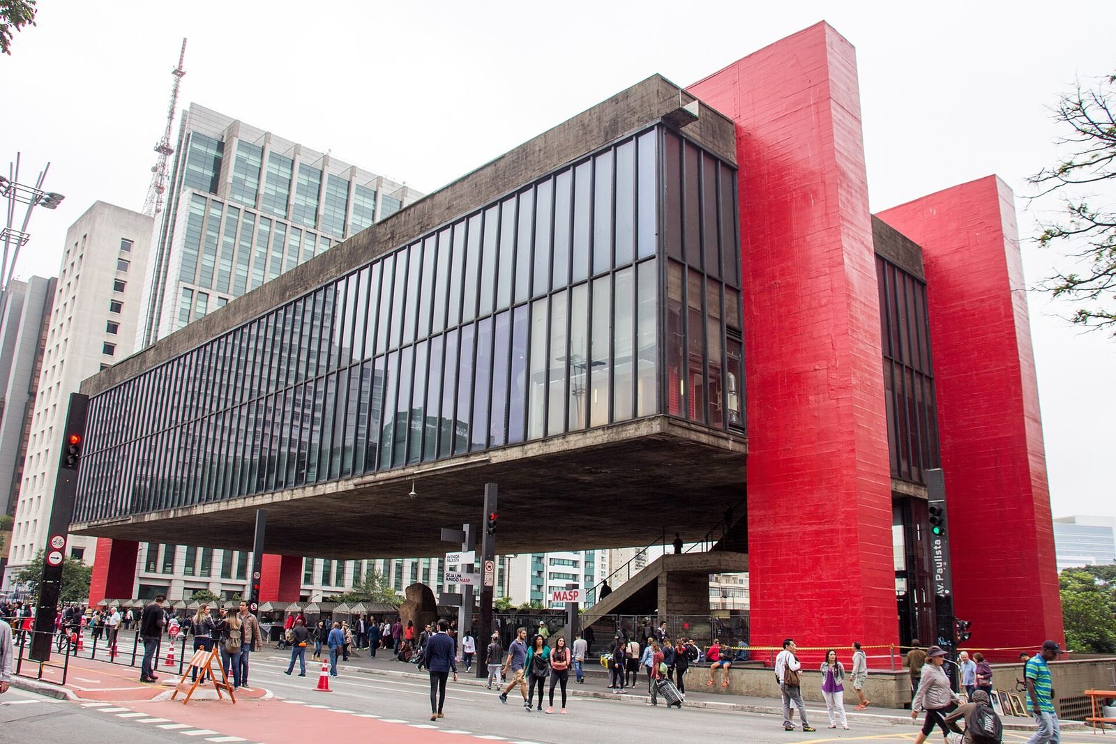 Box of dark glass and concrete suspended over the sidewalk, held between two large red portal frames, with pedestrians walking underneath and tall buildings in the background