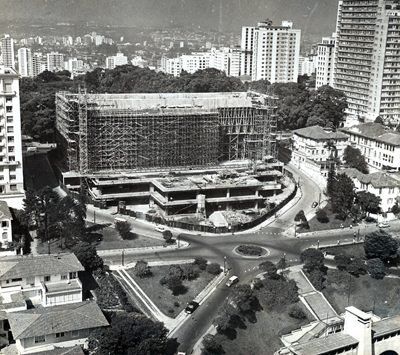 Fotografia aérea em preto e branco: um bloco retangular coberto de andaimes sobre uma base de concreto, ao lado de uma rotatória, cercado de árvores e prédios