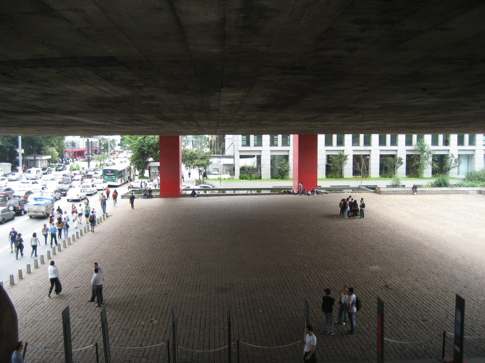 Vista de baixo de uma enorme laje de concreto escuro sobre um piso de paralelepípedos quase vazio, com dois pilares vermelhos ao fundo e a avenida à esquerda