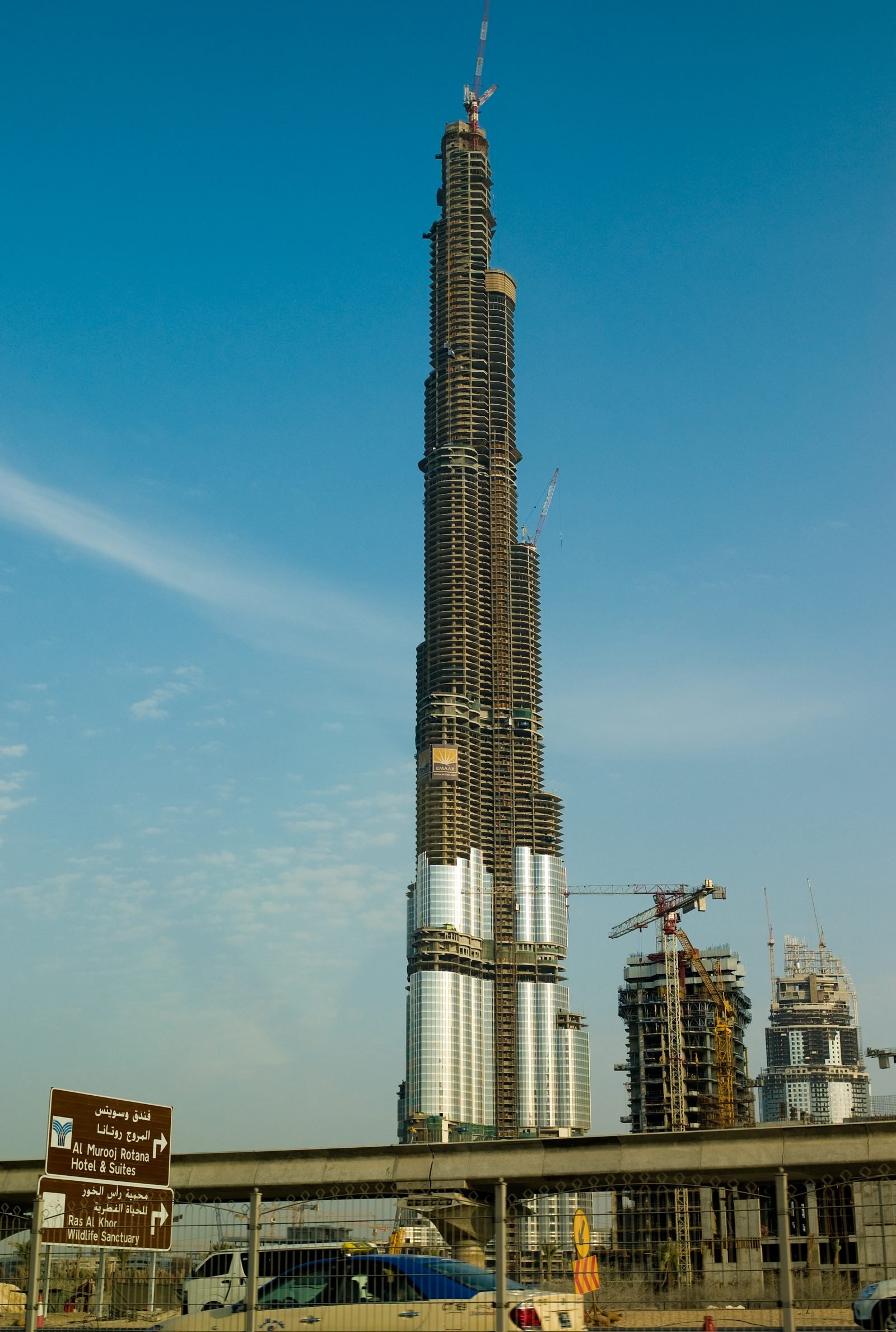 La torre vista desde el suelo contra el cielo azul, con paneles de vidrio plateado solo en la parte de abajo, el concreto a la vista por encima y grúas en lo alto; una señal de tránsito y un viaducto en primer plano