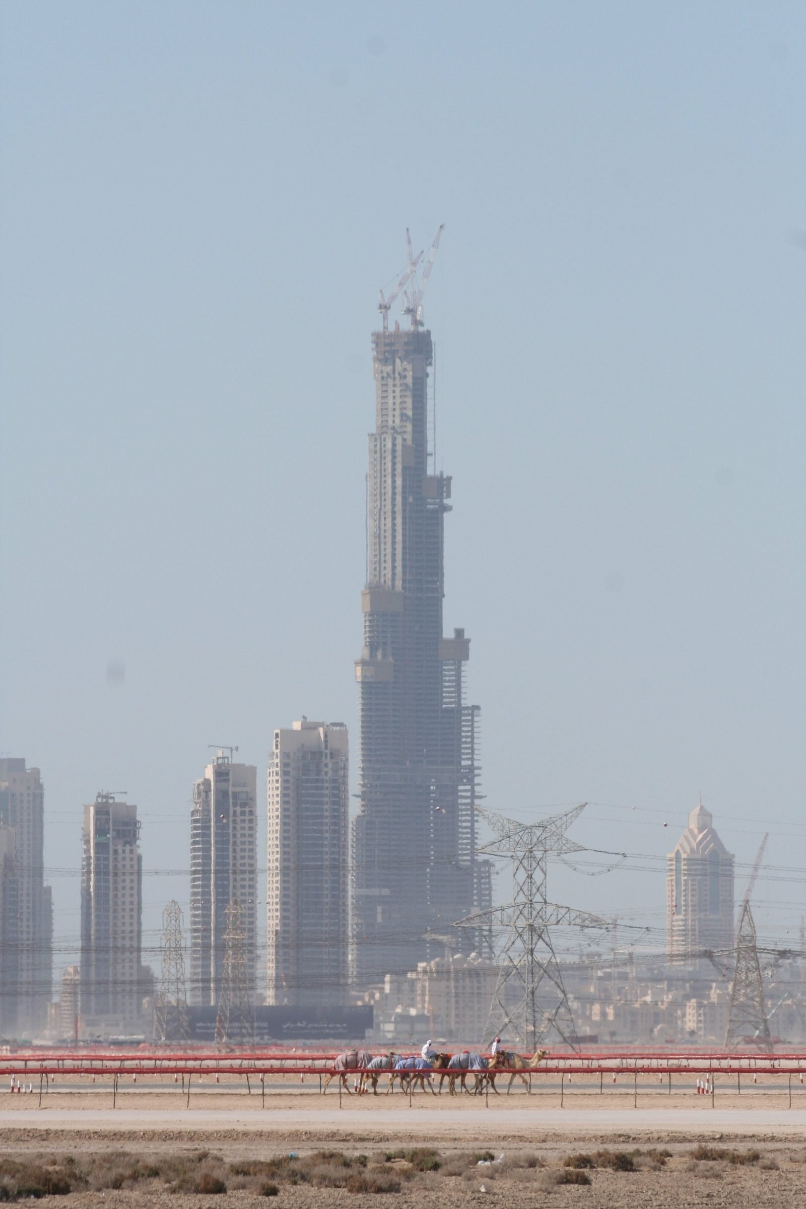 Camellos corriendo en una pista de arena en primer plano, con torres de alta tensión y edificios al fondo y la torre en construcción, con grúas en lo alto, destacándose entre la bruma