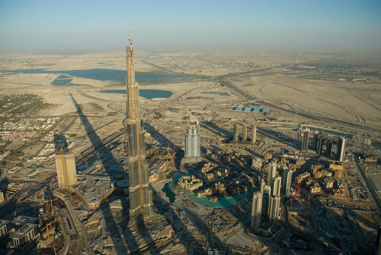 Aerial view of a very tall tower under construction, still without cladding at the top, casting a long shadow over a district of new buildings, building sites and the desert beyond