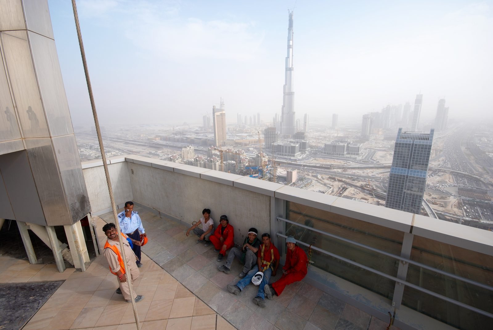 Obreros con overoles naranjas y rojos sentados en el piso de una terraza alta, a la sombra de un murete, con la torre en construcción al fondo entre la bruma