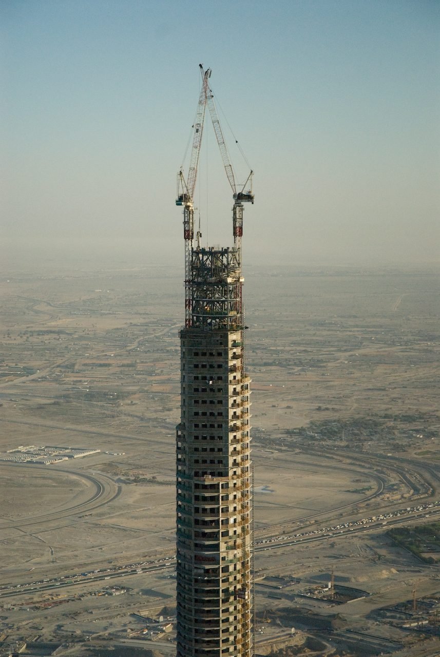 La parte superior de la torre en concreto, todavía en obra, con dos grúas altas y andamios, sobre una llanura de desierto y carreteras vistas desde arriba