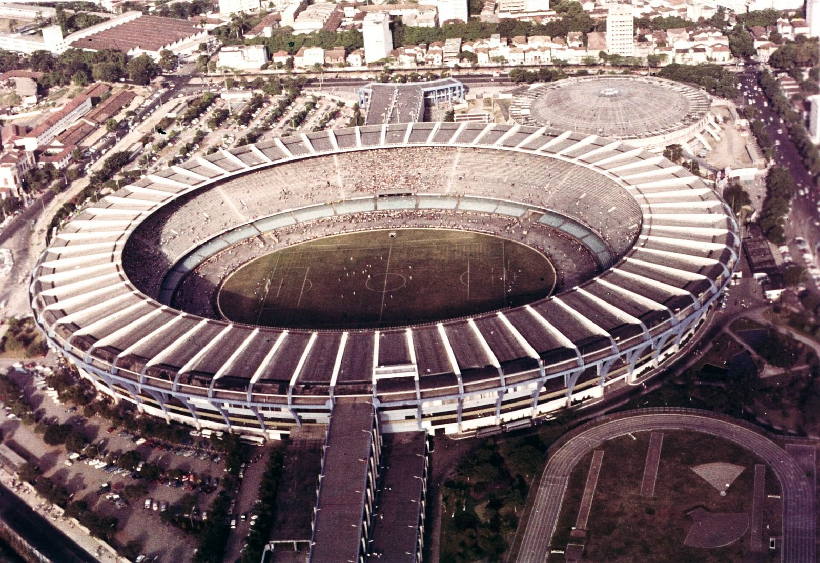 Color aerial view of an oval concrete stadium with the green field at the center, packed stands and a continuous canopy divided into pale segments all the way around; in the background, a round-domed arena and city buildings
