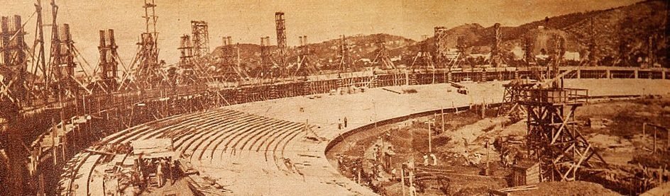 Fotografía antigua en tono sepia, panorámica: graderías de concreto en curva todavía desnudas, rodeadas de altas torres de andamio de madera, con cerros al fondo