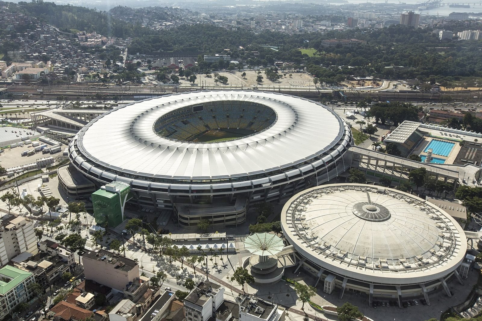 Vista aérea del estadio con una cubierta blanca continua en anillo alrededor del campo, junto al gimnasio de cúpula redonda del Maracanãzinho