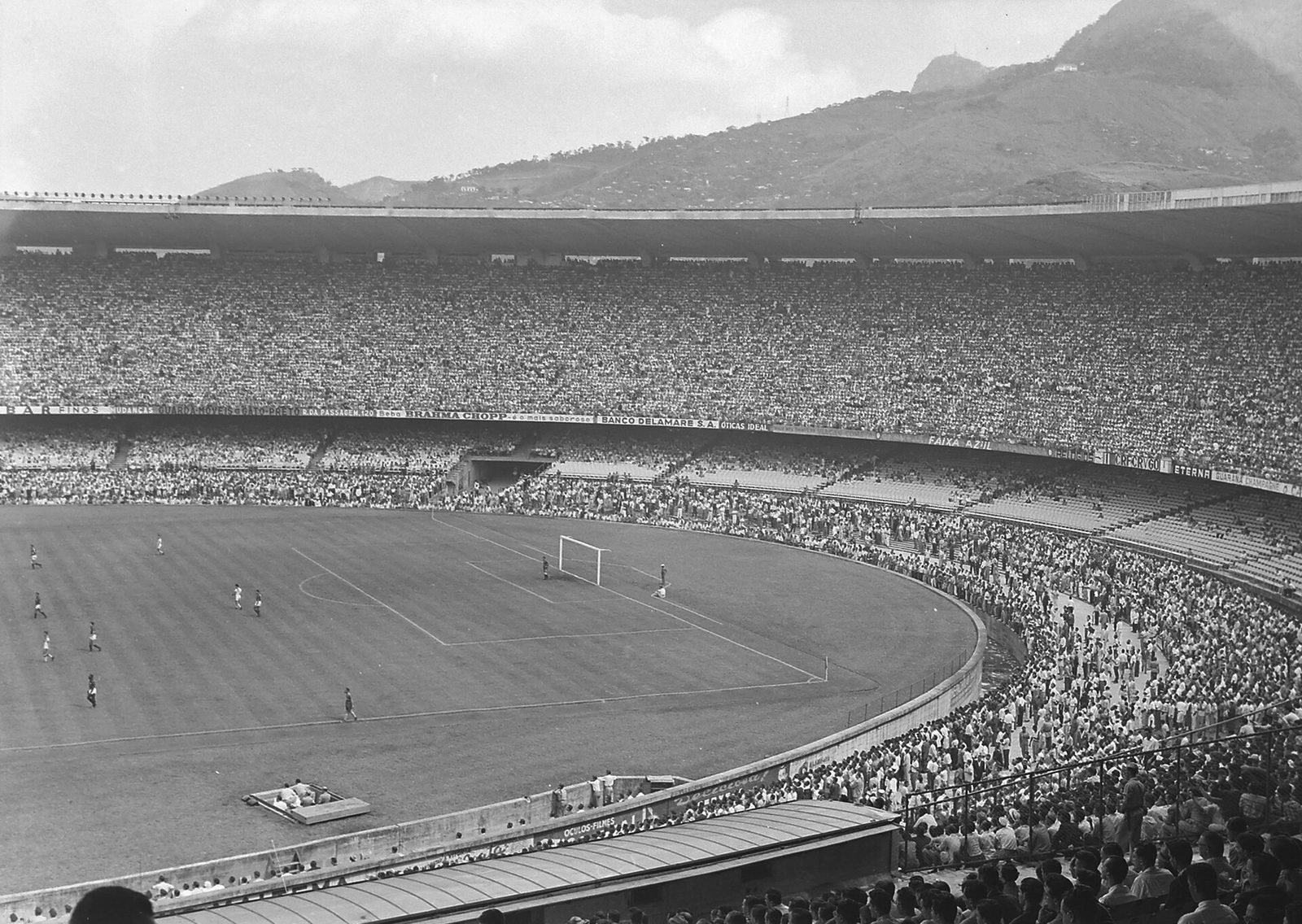 Fotografía en blanco y negro: estadio lleno visto desde lo alto de la gradería, con el campo a la izquierda, jugadores y un arco, y la marquesina oscura bordeando el anillo superior; cerros al fondo
